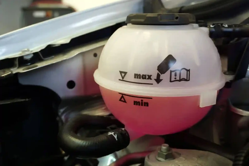 A close-up of a cars coolant reservoir shows pink fluid near the min level marking, with max and min level indicators and a book symbol on the translucent container.