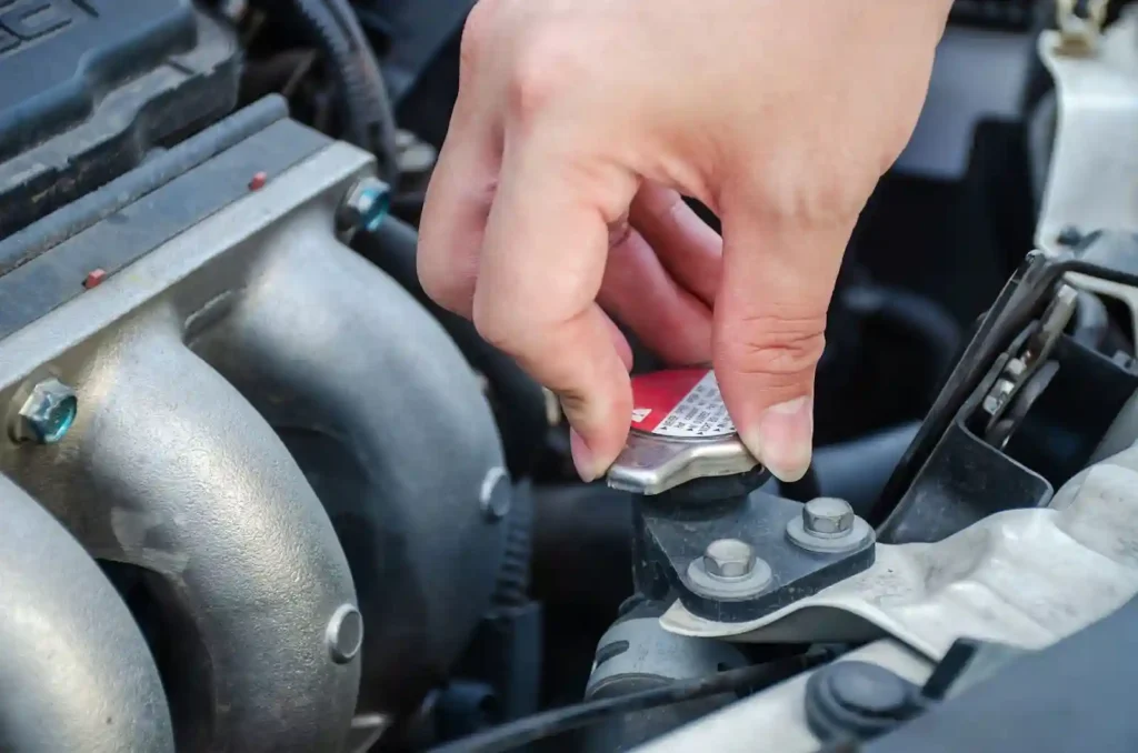 A hand is opening a car radiator cap under the hood, showing part of the engine and radiator components.